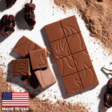 Flamethrower Candy's Ghost Pepper Chocolate bar, unwrapped, with a pattern, surrounded by cocoa powder and dried fruits on a white background, with 'Made in USA' label.