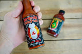 A 142g bottle of 'Da Bomb Evolution' hot sauce held in the hand. A bottle of Da Bomb Beyond Insanity lays on the wooden floor below  - buy online Australia | Blonde Chilli