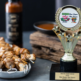 A bottle of Grumpy Gary's The Ripper Hot Sauce in the background. A trophy stands in the foreground beside a plate of chicken skewers.