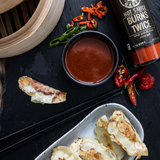 Gyoza with Grumpy Gary's Burns Twice dipping sauce in a bowl, and a bottle of Grumpy Gary's Burns Twice hot sauce on a dark table