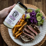 A jar of Grumpy Gary's Gar-Lick Dip with a plate of meat, chips and salad in the background.