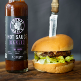 Burger with a bottle of Grumpy Gary's Garlic Hot Sauce on a wooden surface.