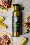 Bottle of Grumpy Gary's Jalapeno Hot Sauce surrounded by tacos and lime wedges on a gray surface