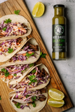Tacos with coleslaw and lime wedges on a wooden board with a bottle of Grumpy Gary's Jalapeno hot sauce.