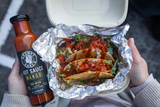 Open container with tacos and a bottle of Grumpy Gary's Mango hot sauce held by a person.