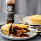 A cut meat pie on a plate with a bottle of Grumpy Gary's Smokey Hot Sauce in the background.
