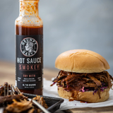 Grumpy Gary's Smokey hot sauce bottle next to a pulled pork sandwich on a wooden surface.