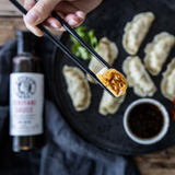 Person holding chopsticks with a dumpling over a plate of dumplings and a bottle of Grumpy Gary's Teriyaki dipping sauce.