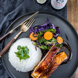 Plated dish with salmon, vegetables, and rice on a dark plate with a bottle of Grumpy Gary's Teriyaki sauce in the background.