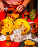 Table with food and drinks including burgers, fries, and Hot Ones hot sauce bottles.