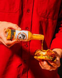Person in a red shirt pouring Piko Peppers hot sauce from a bottle onto a sandwich.