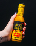 Hand holding a bottle of Hot Ones Tropical Amarillo hot sauce against a black background