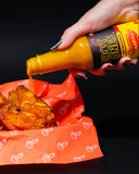 Hot sauce being poured onto fried chicken on a red paper with white patterns.