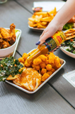 Person dipping cauliflower into a bottle of Hot Ones Tropical Amarillo hot sauce on a table with other food items.