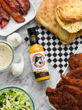A 148ml bottle of Microsaucerie Piko Peppers Garlic Delight Hot Sauce on a marble table surrounded by bacon, bread, schnitzels, and garlic.