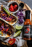 Spicy tacos with purple cabbage, red peppers, and a bottle of 13 Angry Scorpions' The Notorious hot sauce on a wooden board.