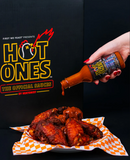 Hand pouring Hot Ones sauce onto fried chicken with the Hot Ones logo in the background.