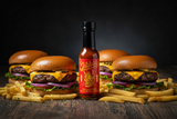 A bottle of Jibba's Jalapeno Black Garlic hot sauce stands on a table amongst 4 burgers and fries.