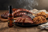 A bottle of Jibba's Reaper BBQ Hot Sauce stands in the foreground of an American BBQ feast.