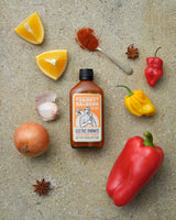 Danny Balboa's ELECTRIC DYNAMITE - Capsicum, Orange and Habanero Hot Sauce, lying on a table surrounded by fresh ingredients as found in this hot sauce