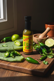 A bottle of Jibbas Nopale Verde on a wooden table amidst some green jalapeno, nopales and lime.