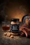 Uncle Mungo's Brown Sugar Bourbon  Dry Rub & Seasoning on a wooden table surrounded by oak barrels, a glass of bourbon, brown sugar, and beef ribs.